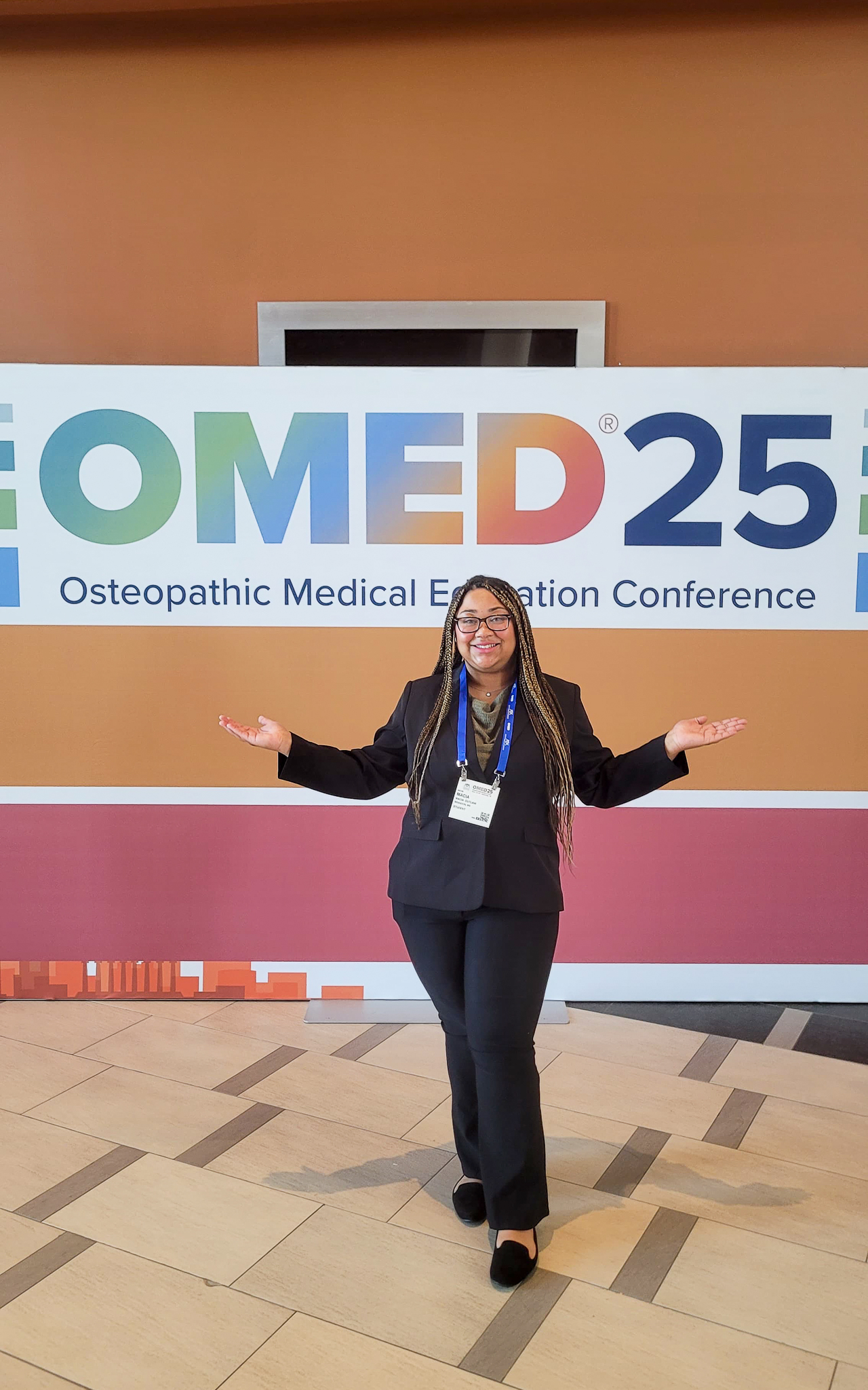 BUCOM student poses in front of the OMED 2025 conference sign in Nashville, smiling with arms open in excitement.