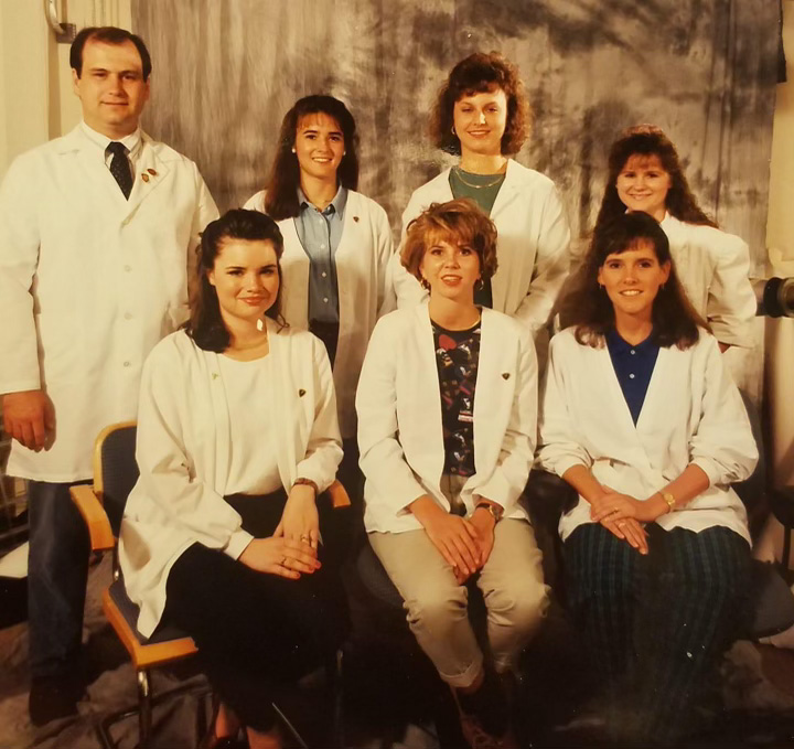 7 sonography students wearing white lab coats posing for a photo. Three students are sitting down while four are standing behind them