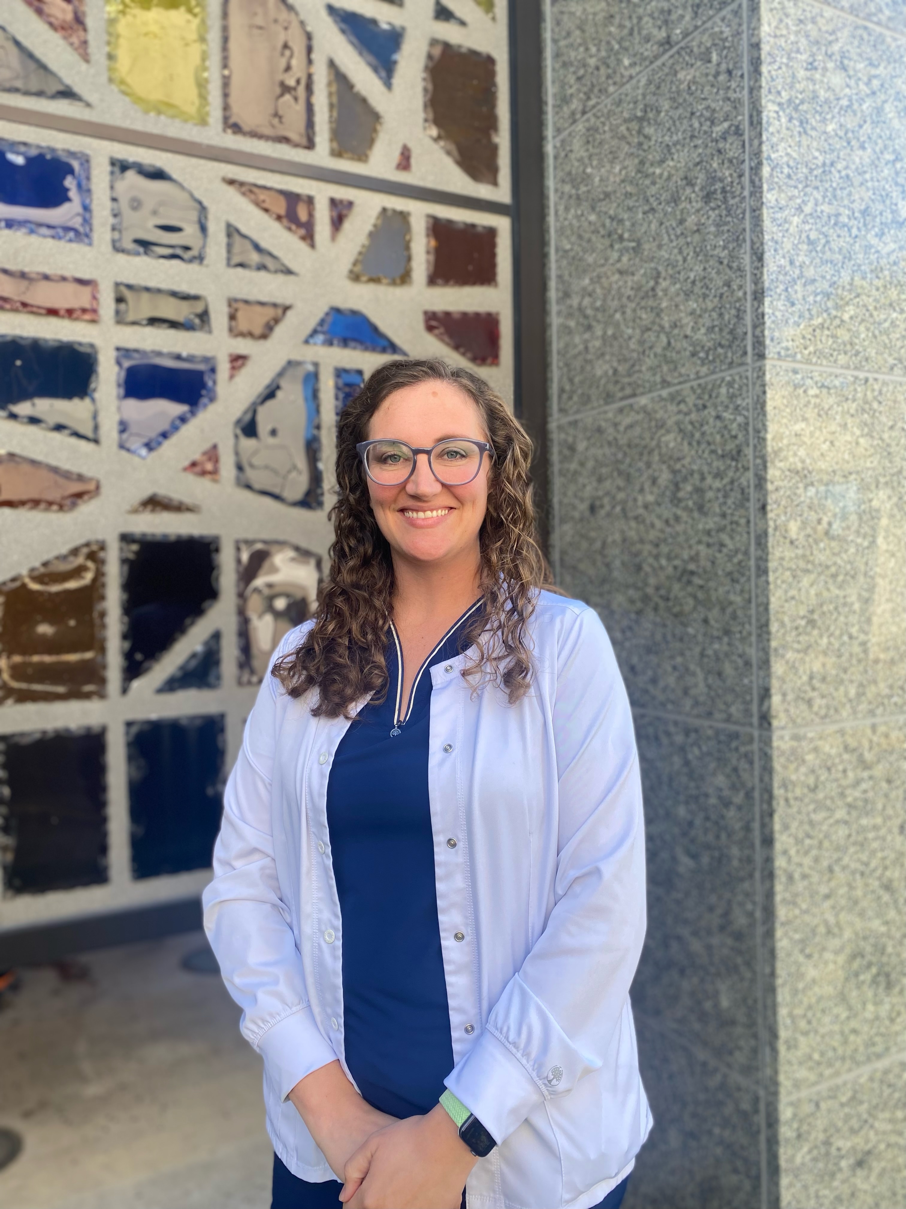 April Smith with her back on a wall smiling at the camera in blue scrubs and white lab coat