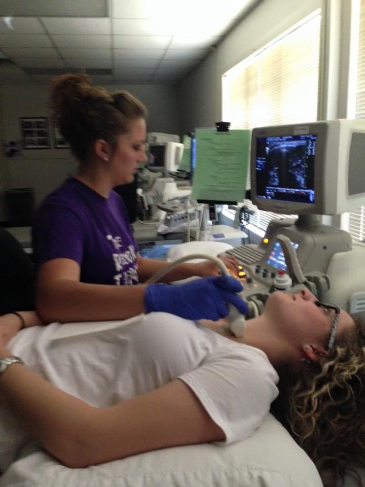 Sonography student with a purple shirt on performing a checkup on a classmate who is laying down