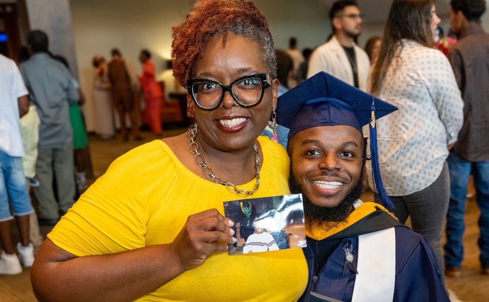 Photo of student wearing a cap and gown and his mother sitting on top of him with a yellow dress on