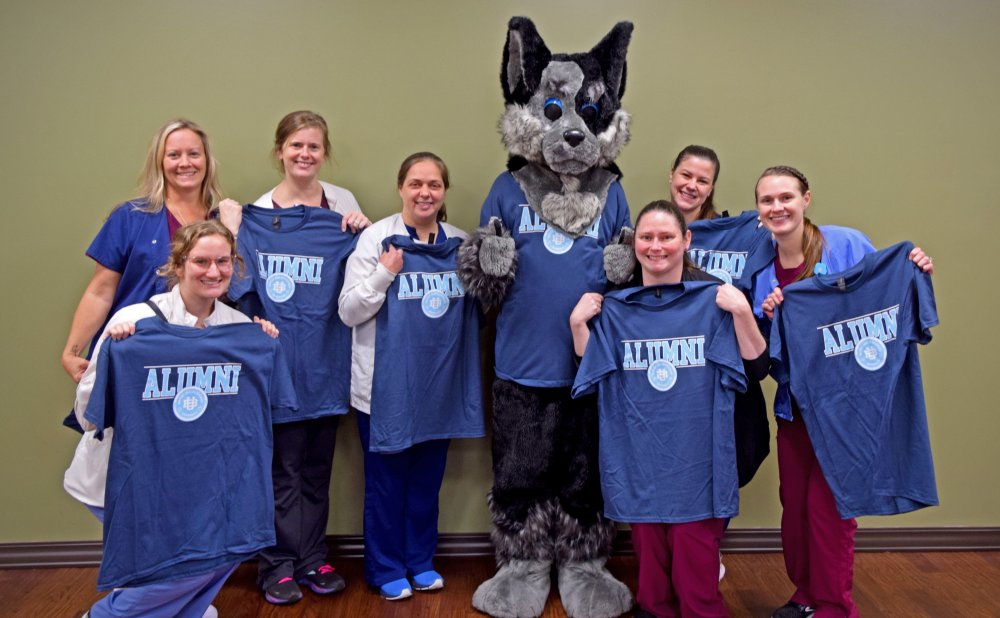 8 DMS student alumni holding blue alumni shirts with light blue lettering posing with Hartley the dog mascot of the university. Six have on red scrubs and one has on blue scrubs