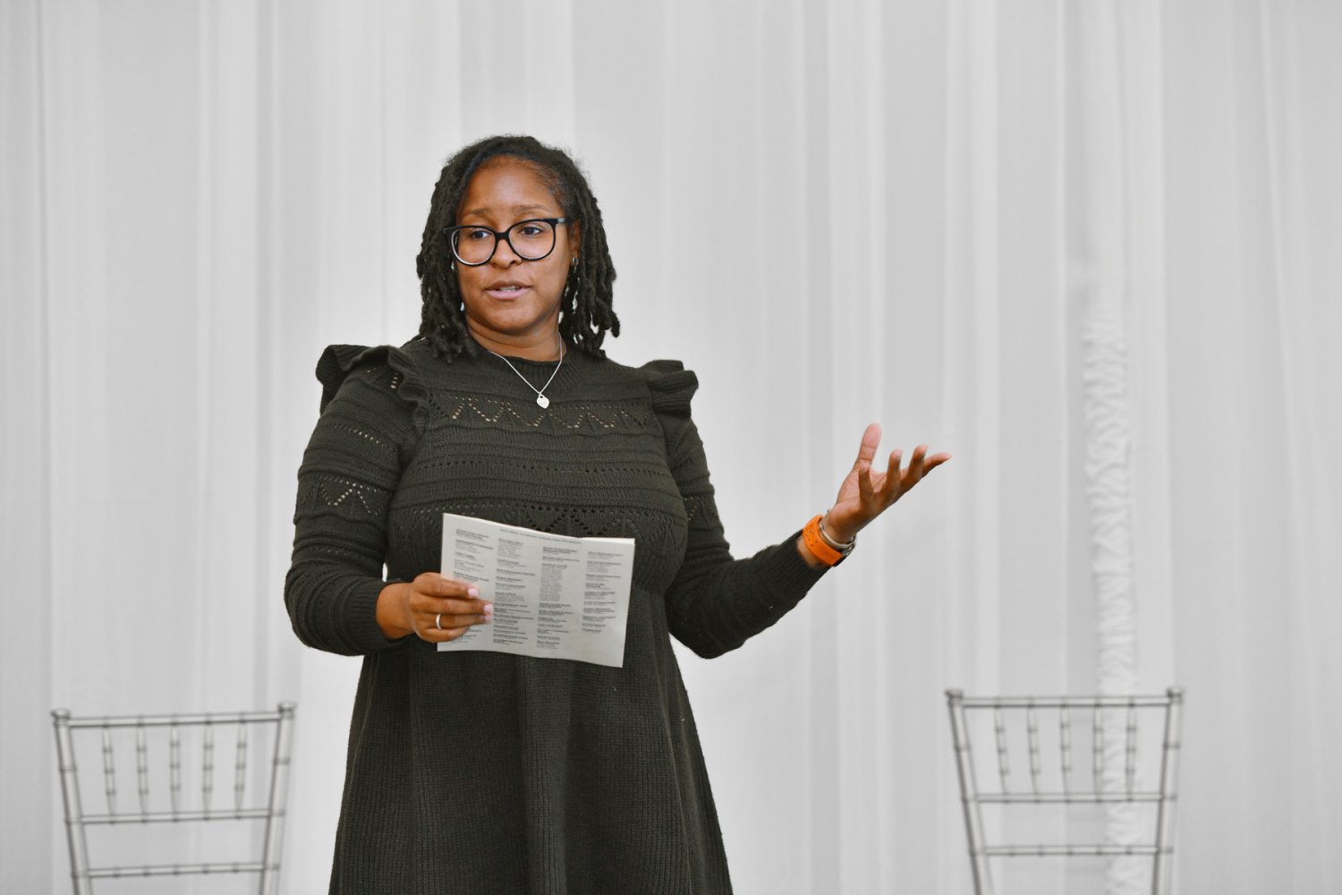 A woman wearing glasses and a dark dress gestures while speaking in front of a white curtain.