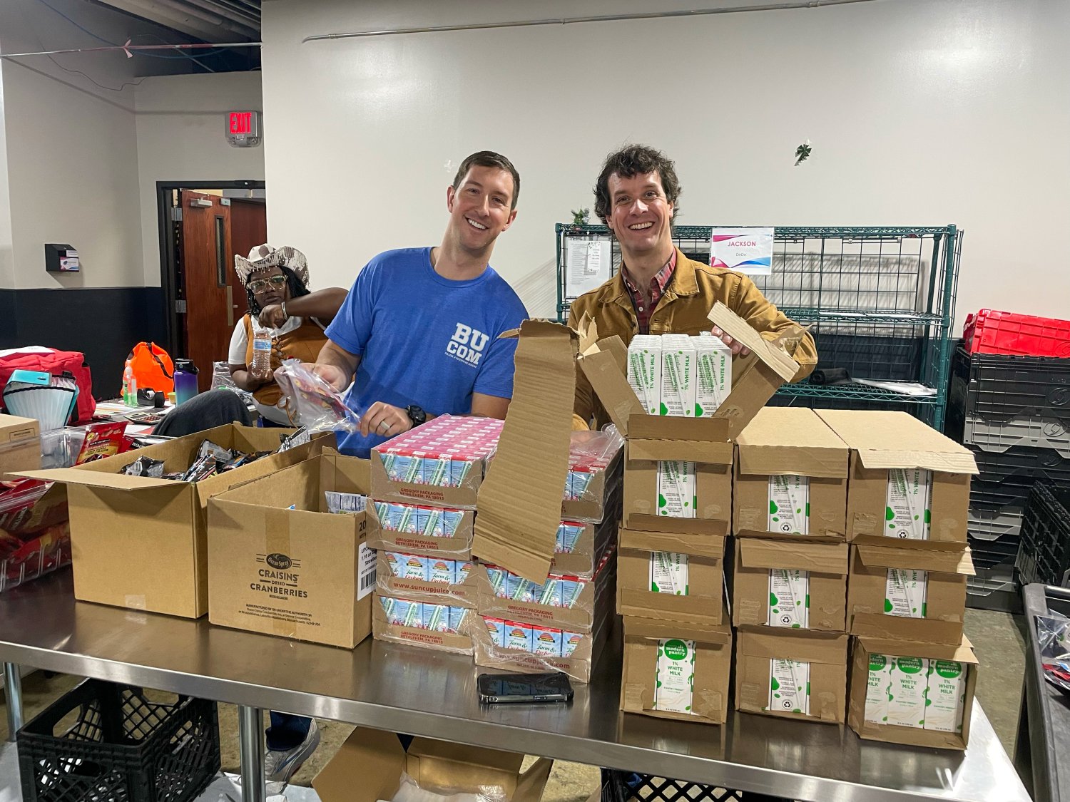 two BUCOM students packaging items taking a photo behind boxes 